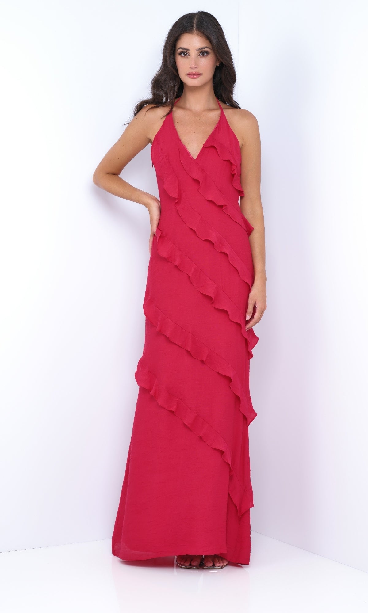A woman stands against a white background wearing the Remi by Velvi, a V-neck halter ruffled long prom dress in vibrant red with elegant diagonal ruffle detailing.